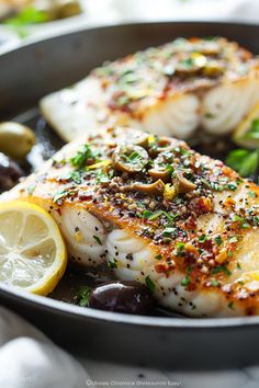 two fish in a skillet with olives and lemon slices on the side for garnish