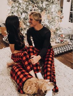 two people sitting on the floor next to a christmas tree with a dog in front of them