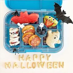 a plastic lunch box filled with halloween food and decorations, including cookies, pretzels, fruit, and bats