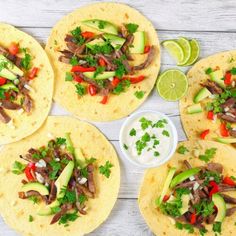 four tortillas topped with meat, vegetables and avocado on a white wooden table