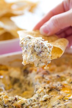 a hand holding a tortilla chip over a casserole dish filled with chicken and cheese