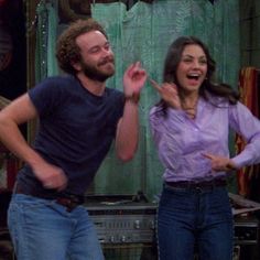 a man and woman standing next to each other in front of a green curtain with their hands up