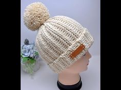 a white knitted hat with a leather patch on the front and a pom - pom at the top