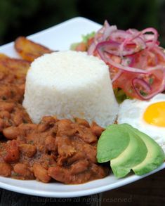 a white plate topped with beans and rice next to vegetables on a wooden table top