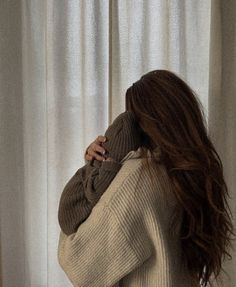 a woman with long brown hair standing in front of a window wearing a sweater and jeans