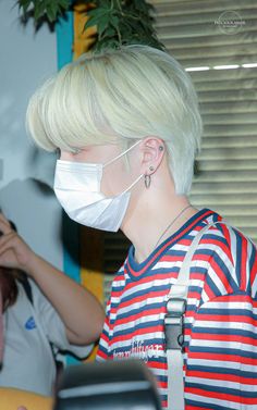 a woman with blonde hair wearing a face mask and looking at her cell phone while another person in the background looks on