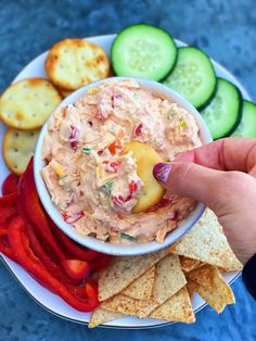 a hand holding a cracker over a bowl of dip surrounded by chips and cucumbers