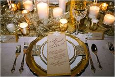 the table is set with gold and white plates, silverware, candles and napkins