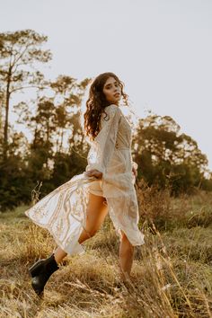 a woman in a white dress and black boots is walking through the grass with trees behind her