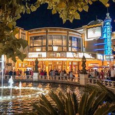 people are walking around in front of a building with a fountain at the center and lights on