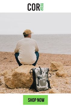 a man sitting on top of a rock next to the ocean wearing a hat and sweater