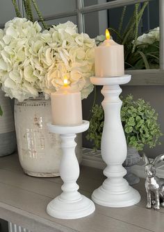 two white candles sitting on top of a wooden table next to flowers and a mirror