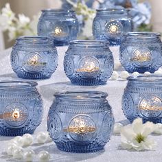 several blue glass jars with lit candles in them on a table covered with white flowers