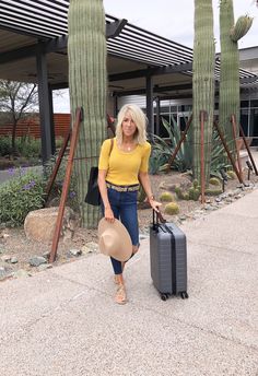 a woman with a hat and suitcase walking down the street in front of cactus trees