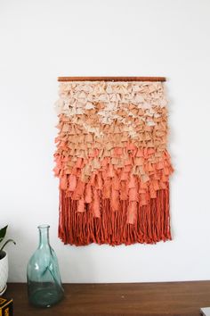 a wall hanging made out of orange and white fabric with fringes on it next to a vase