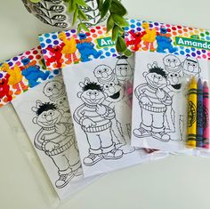 three coloring books and two crayons are sitting on a table next to a potted plant