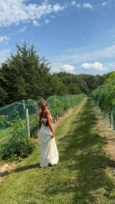 a woman in a long white dress walking down a path between rows of vines and trees