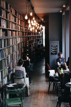 people sitting at tables in a library with lots of bookshelves and lamps hanging from the ceiling