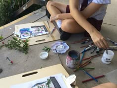 a woman sitting on the ground painting with paintbrushes and other art supplies next to her