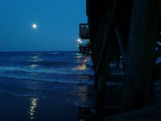 the moon is setting over the ocean with waves crashing in front of it and a pier