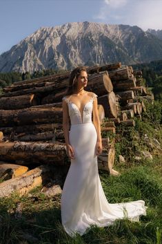 a woman in a wedding dress standing next to some logs and grass with mountains in the background