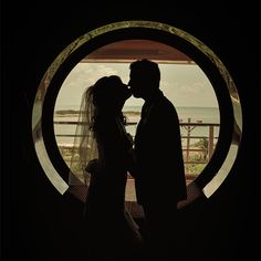 a bride and groom kissing in front of a circular window with the ocean behind them