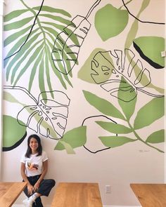 a woman sitting on top of a wooden bench in front of a wall with leaves painted on it