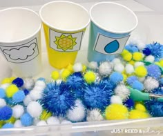 three cups with pom - poms are sitting in a plastic container filled with blue and yellow balls