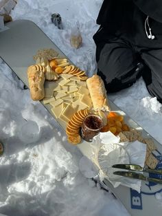 a snowboard with cheese and crackers on it sitting in the snow next to other food