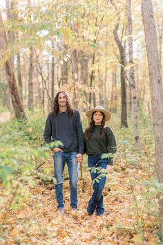 two people standing in the woods with leaves on the ground and one person wearing a hat