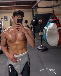 a shirtless man taking a selfie in a gym with boxing gloves and punching bag