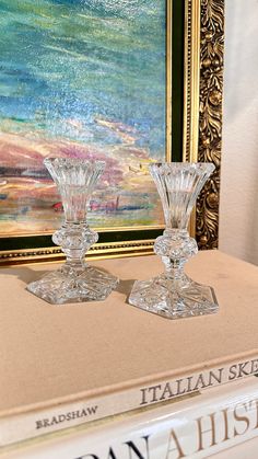 two crystal candlesticks sitting on top of a book in front of a painting