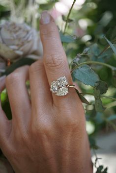 a close up of a person's hand with a ring on their finger and flowers in the background