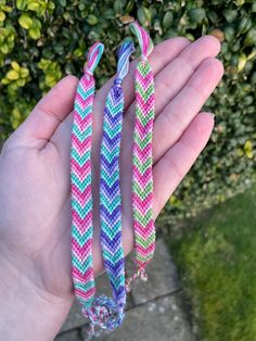 three colorful bracelets are being held in someone's hand with green bushes behind them