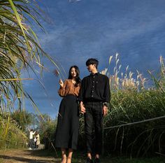 a man and woman standing next to each other in front of some tall grass on a sunny day