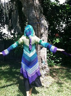 a woman wearing a colorful crocheted dress and holding her arms out in front of a tree