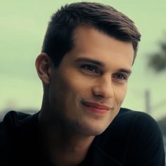 a close up of a person wearing a black shirt and smiling at the camera with trees in the background