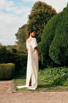 a woman in a white dress is standing on the side of a road with trees and bushes behind her