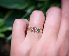 a person's hand holding a ring with three different colored stones on it and green leaves in the background