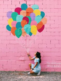 a woman sitting in front of a pink brick wall holding onto a bunch of balloons