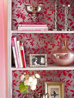 a book shelf with vases, books and pictures on it in front of a wallpapered background