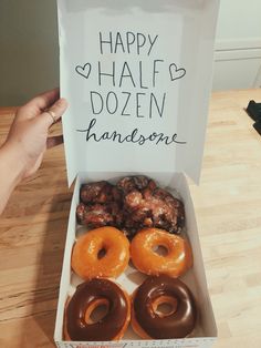 a box filled with lots of donuts on top of a wooden table next to a handwritten sign