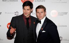 two men standing next to each other in front of a red carpet and white wall