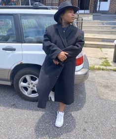 a woman standing in front of a car wearing a coat and white tennis shoes with her feet on the ground