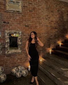 a woman in a black dress standing next to some stairs with candles on the wall