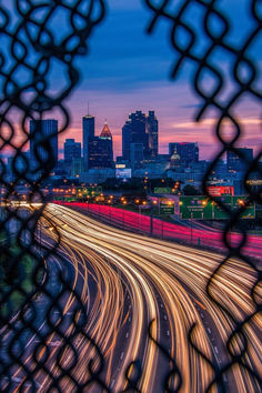 the city skyline is seen through a chain link fence at night with light streaks in the foreground