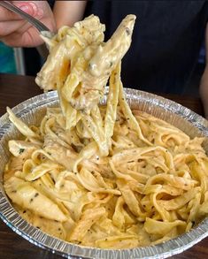 a person holding a fork full of pasta in a tin foil pan on a wooden table