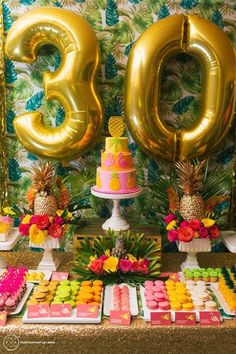 a table with pineapples, pink and yellow desserts and large gold balloons