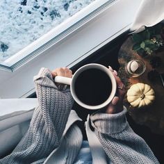 a person holding a cup of coffee in their hands while looking out the window at snow and pumpkins