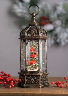 a christmas tree in a glass and metal lantern with red berries on the table next to it